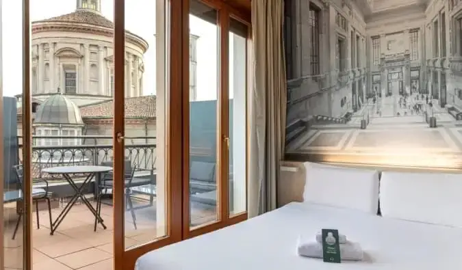 Een tweepersoonsbed in een hotelkamer met een open deur die leidt naar een terras met uitzicht op een basiliek in het B&B Hotel Milano Sant