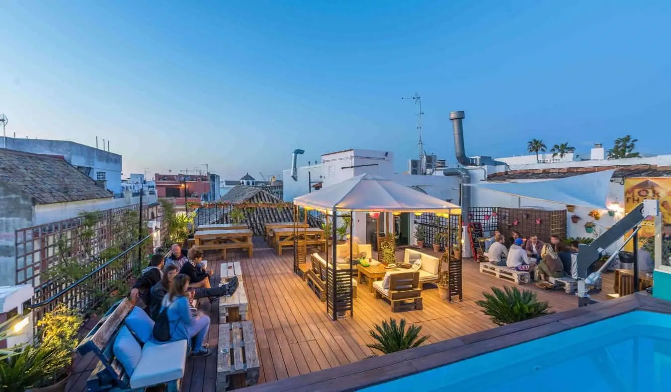 Gente descansando en una gran terraza en la azotea con la esquina de una piscina en primer plano y el paisaje urbano de Sevilla, España, al fondo