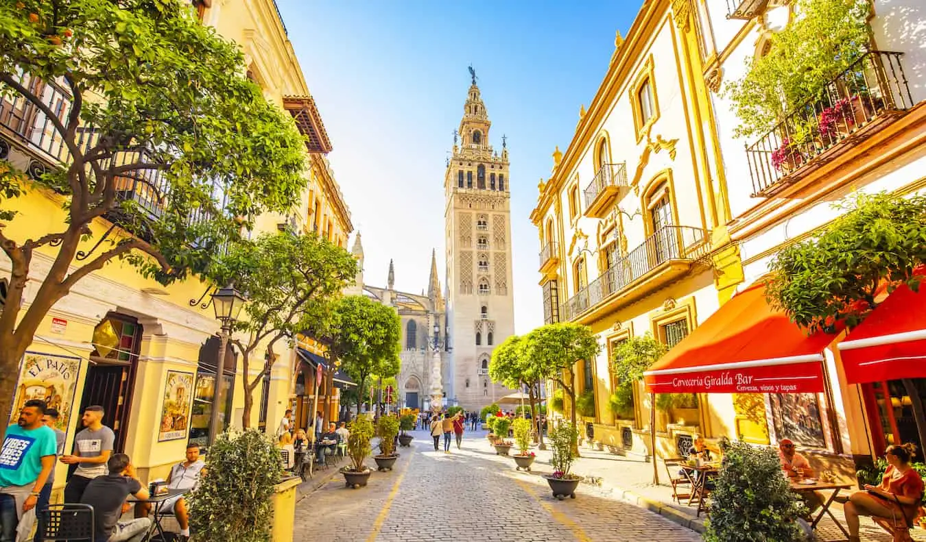 Gente caminando por una calle estrecha y tranquila en la soleada Sevilla, España, con la torre de una iglesia en la distancia