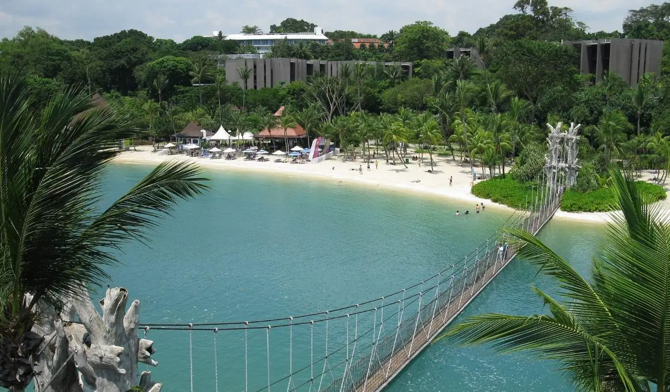 En hengende hengebro for fotgjengere som fører til en sandstrand med palmetrær og små hytter i Sentosa, Singapore