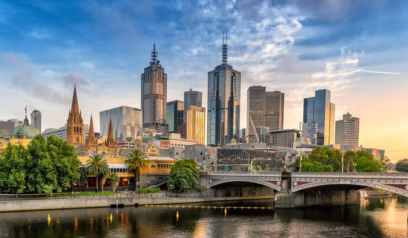 Den ruvende skyline av Melbourne, Australia med trær og en bro i forgrunnen nær elven