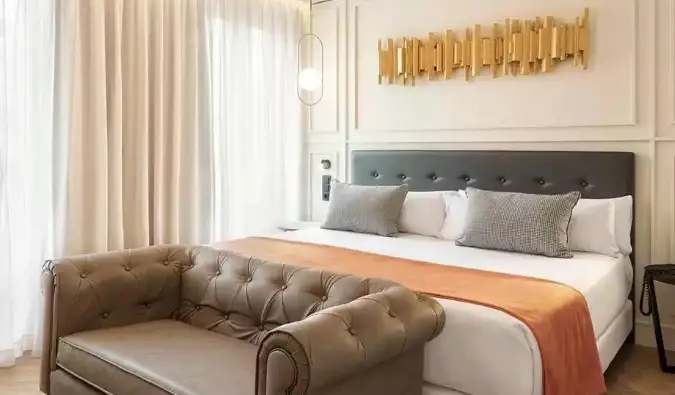 Un pezzo di arredamento dorato è appeso sopra un letto matrimoniale con un divanetto ai piedi del letto presso l'Hotel Catalogna Goya a Madrid, Spagna