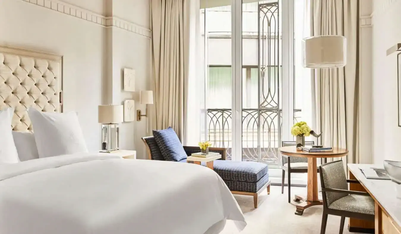 Ein luxuriöses cremefarbenes Hotelzimmer mit einem großen Bett, einer blauen Chaiselongue am offenen Fenster und einem Schreibtisch im Four Seasons in Madrid, Spanien