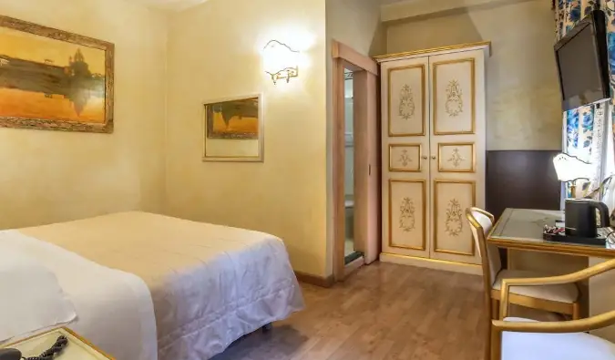 Um quarto de hotel com uma cama de casal simples, uma escrivaninha, pinturas de Florença nas paredes amarelas suaves e um guarda-roupa dourado no canto do Hotel Alba Palace em Florença, Itália