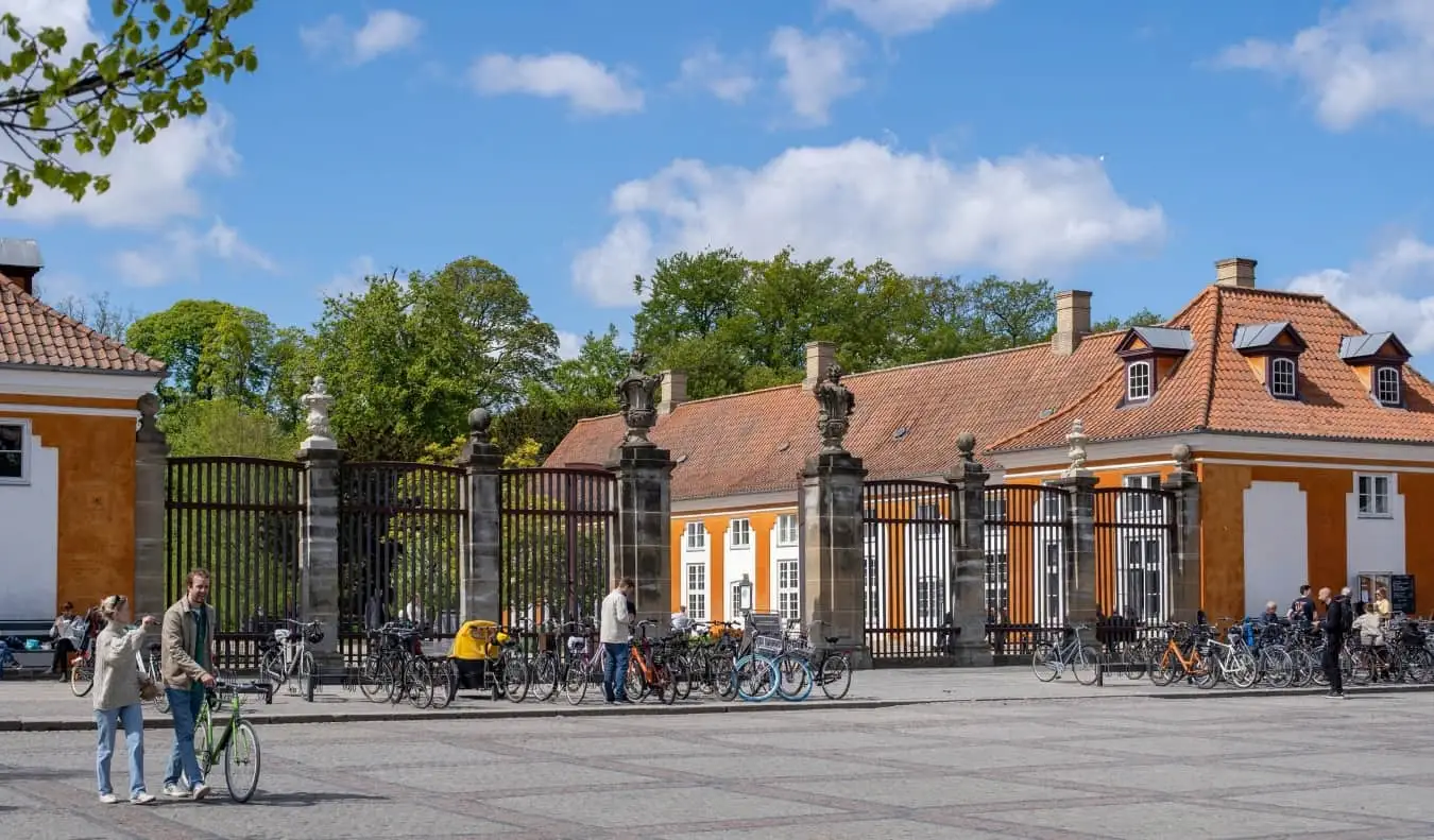 Folk, der går forbi en stor gated indgang til en træfyldt park, flankeret af historiske orange bygninger i København, Danmark