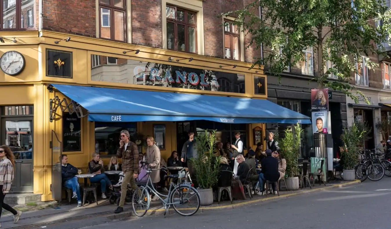 Orang ramai berjalan di tepi orang yang duduk di kafe luar di jalan berdaun di kejiranan Vesterbro di Copenhagen, Denmark