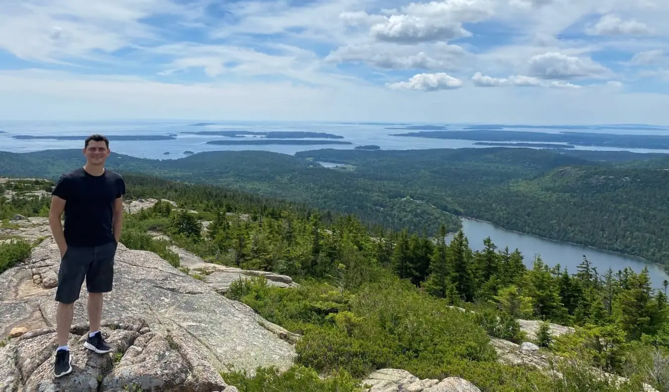 Matt står på toppen av et fjell med en frodig skjærgård bak seg i Maine, USA