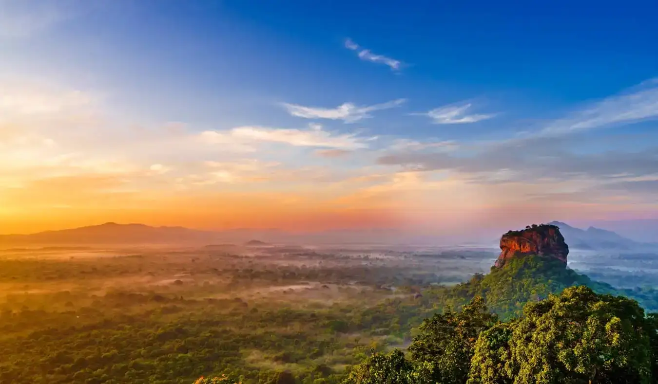 Soluppgång över ett vidsträckt frodigt landskap med Sigiriya-klippan, en stor klippformation, som reser sig över träden i Sri Lanka