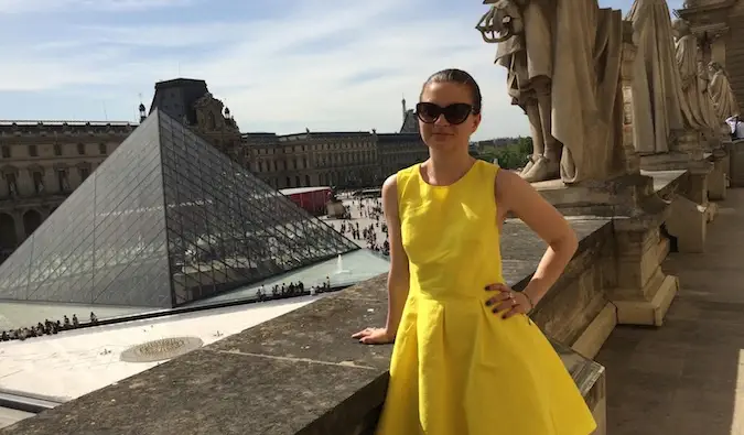 En kvinnlig ensamresenär i en gul klänning poserar framför Louvren, Paris