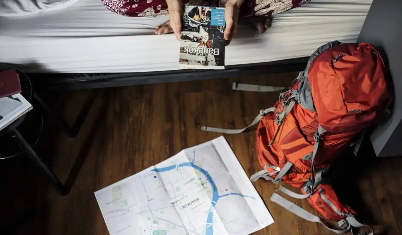 Uma pessoa sentada em um beliche em um albergue segurando um mapa de Bangkok, com a mochila no chão próxima