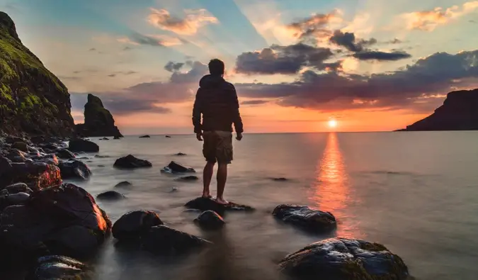 Solo ceļotājs, kas pozē noskaņotam fotoattēlam saulrietā, stāvot pludmalē