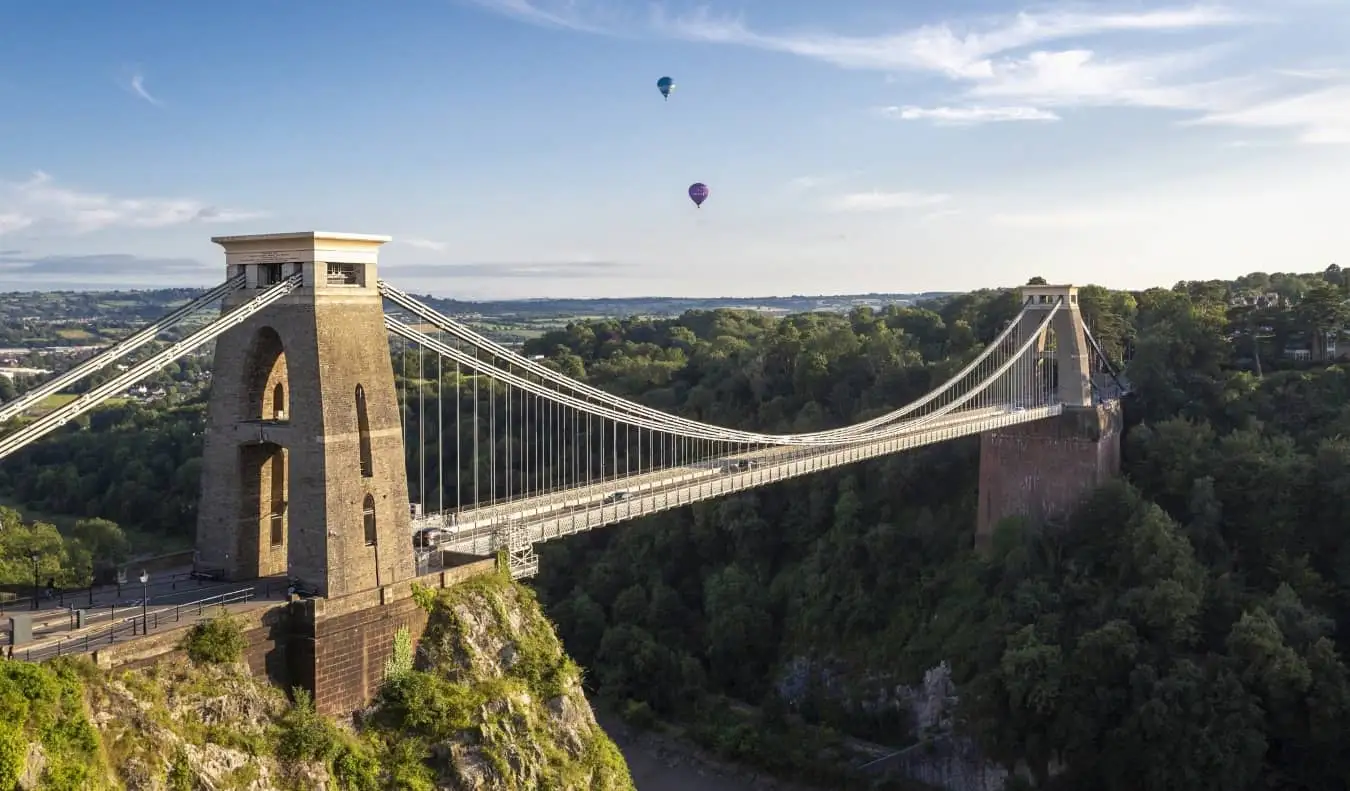 Kijkend naar de Clifton Suspension Bridge die over een diepe kloof snijdt, met een paar heteluchtballonnen op de achtergrond in Bristol, Verenigd Koninkrijk