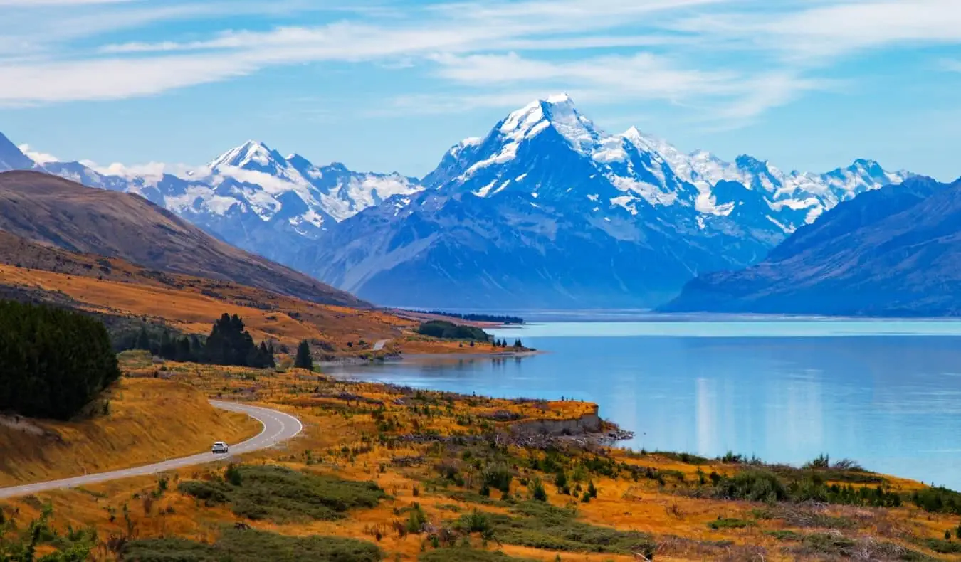 Van roulant sur une route sinueuse avec un lac et des montagnes en arrière-plan en Nouvelle-Zélande.