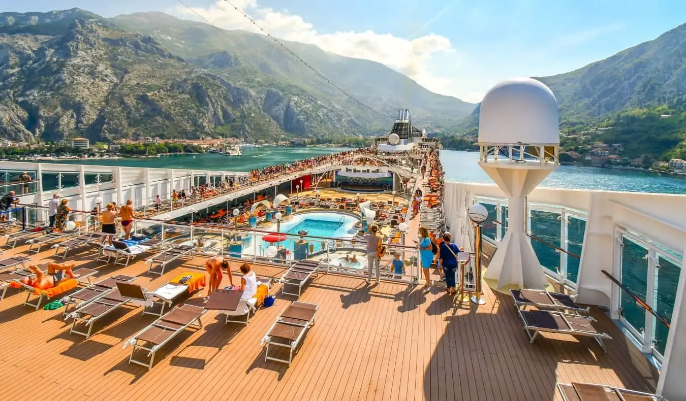 Kreuzfahrtdeck mit Menschen, die auf Stühlen neben einem Pool faulenzen, während das Schiff in die Bucht von Kotor einfährt, mit grünen, sanften Bergen im Hintergrund