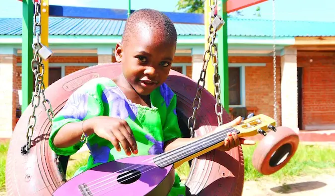 Küçük bir gitar çalan renkli kıyafetler giymiş genç bir Afrikalı çocuk