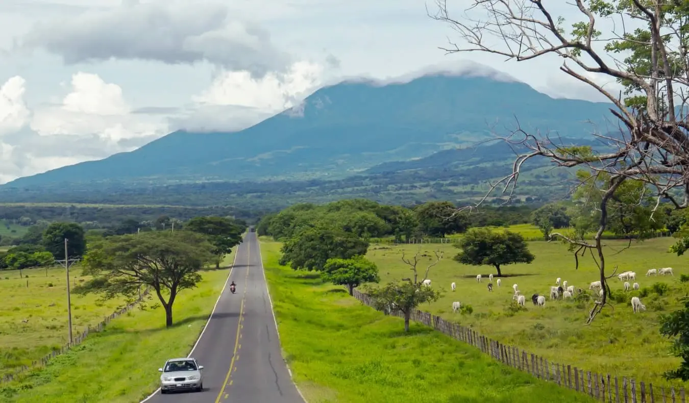 Autos circulando por una carretera en Costa Rica con un volcán al fondo y vacas pastando a los lados de la carretera