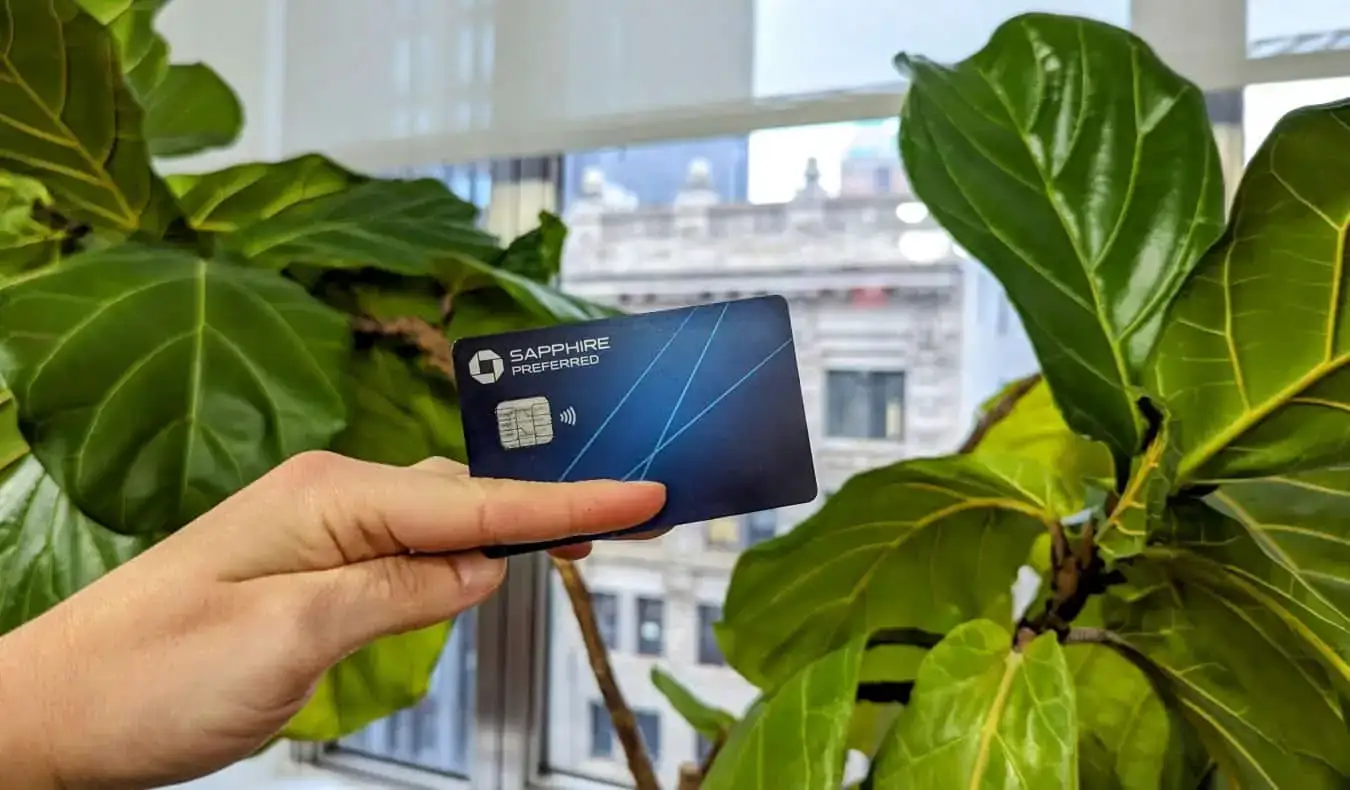 Die Chase Sapphire Preferred Card wird vor einer grünen Pflanze hochgehalten, im Hintergrund sind Gebäude durch das Fenster zu sehen