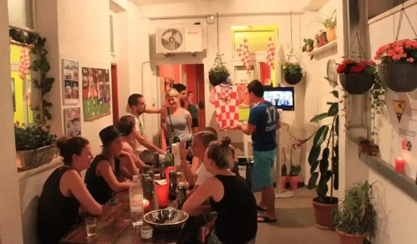 Grupa mugursomnieku, kas sēž pie galda Dubrovnik Backpackers Club koplietošanas telpā, kas ir pilna ar augiem — hostelis Dubrovnikā, Horvātijā.