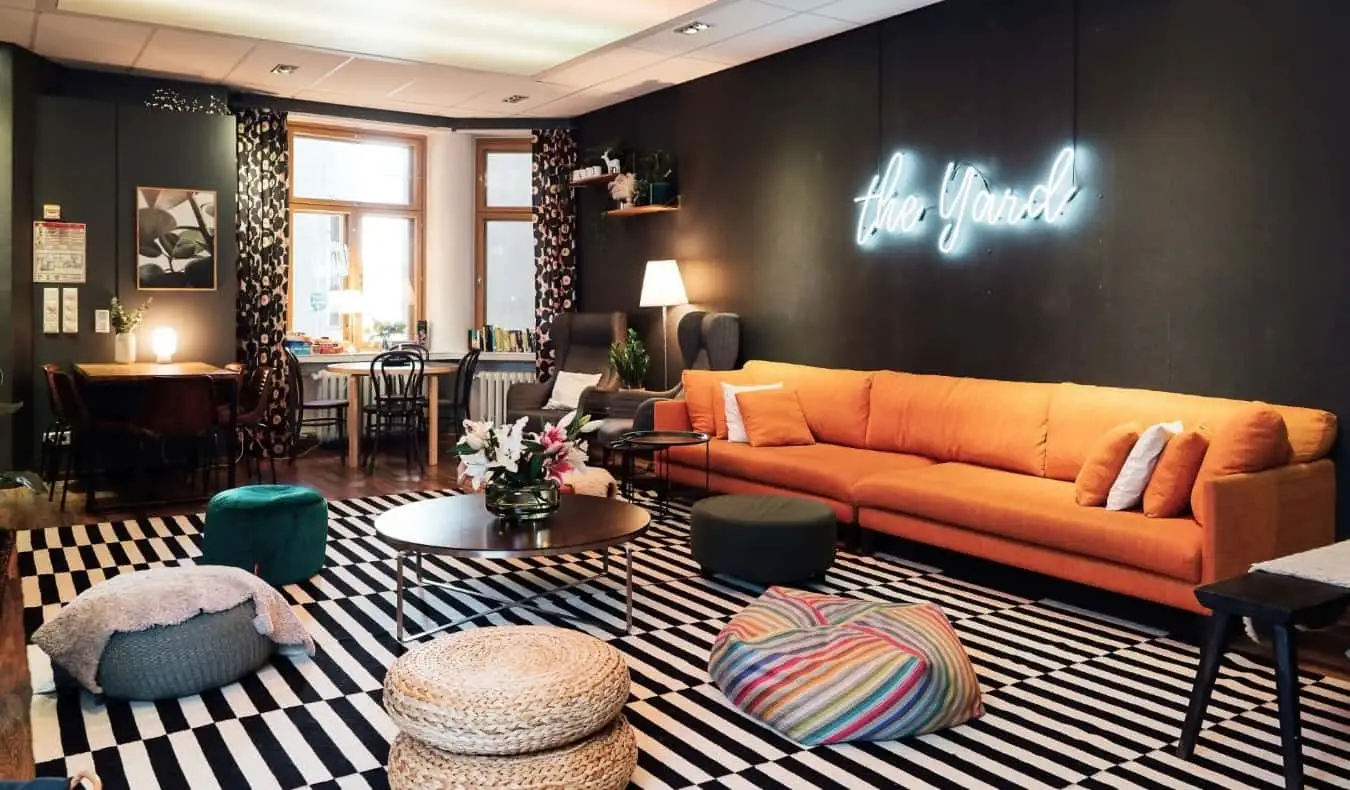 Salle commune chic du Yard Hostel à Helsinki, en Finlande, avec un canapé orange, des bouffées pour s'asseoir et une enseigne au néon indiquant