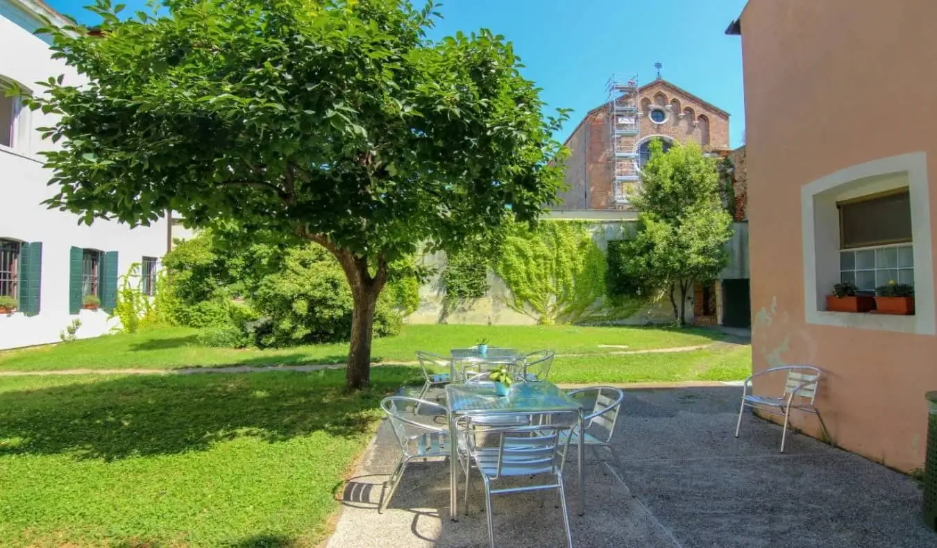 Eingezäunter Hinterhof mit Bäumen, Terrassentischen aus Metall und einer Kirche in der Ferne im Ostello S. Fosca Hostel in Venedig, Italien.