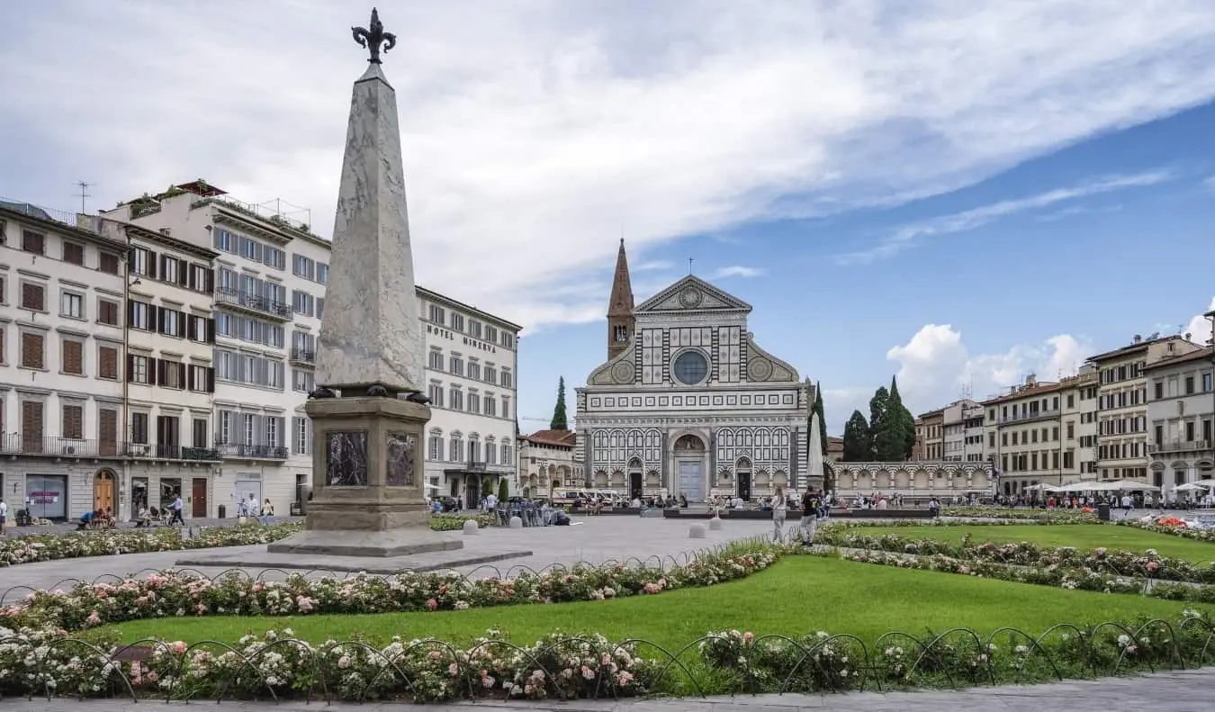 Eine Piazza mit Blumen und Gras rund um ein Denkmal und die bemalte Basilika Santa Maria Novella im Hintergrund in Florenz, Italien