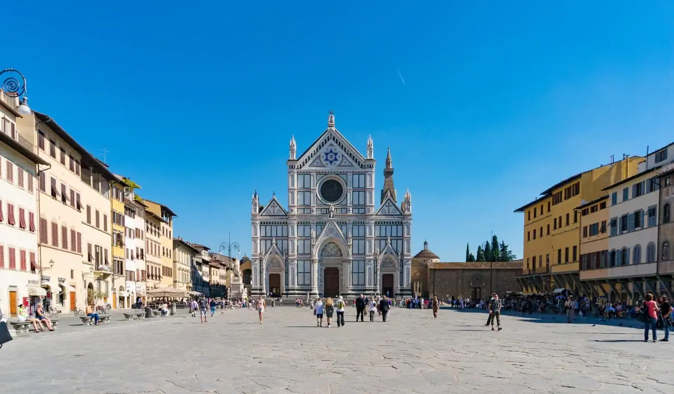 Didelė aikštė, išklota pastatais, o žmonės vaikšto atviroje erdvėje priešais didingai nudažytą Santa Croce baziliką Florencijoje, Italijoje.