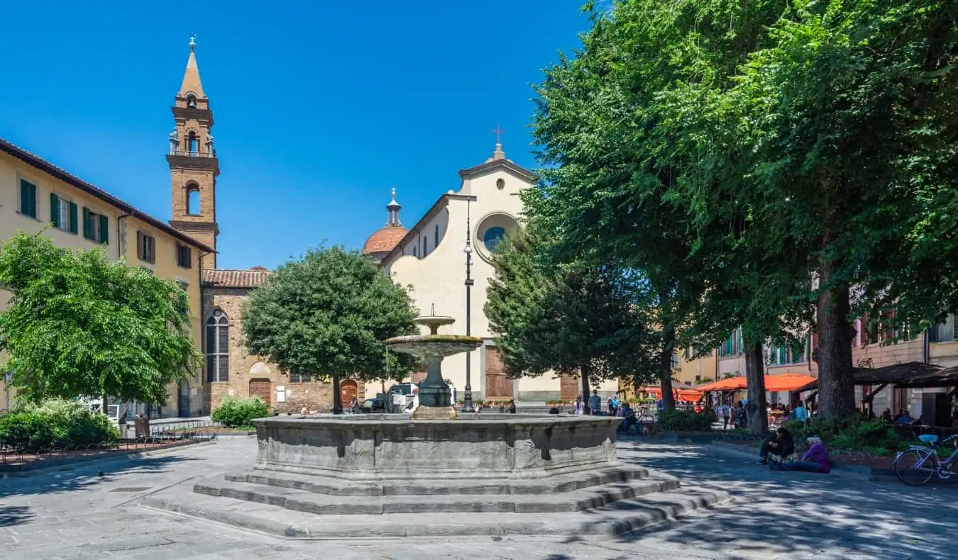 Pessoas sentadas perto de uma fonte na arborizada praça Santo Spirito, na área de Oltrarno, em Florença, Itália