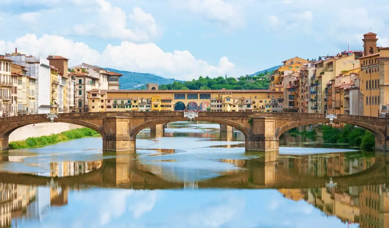 Panoramica del Ponte Vecchio che si estende sul fiume Arno a Firenze, con entrambe le sponde del fiume e il ponte fiancheggiato da edifici gialli