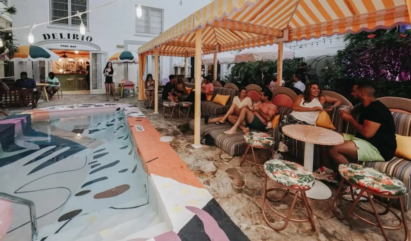 Oameni care stau la mese mici lângă piscina în aer liber cu gresie colorată la Viajero Hostel din Miami, Florida