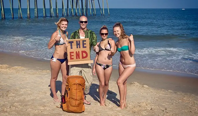 Mann som står på stranden med 3 jenter i bikini og et haikeskilt i papp som sier THE END