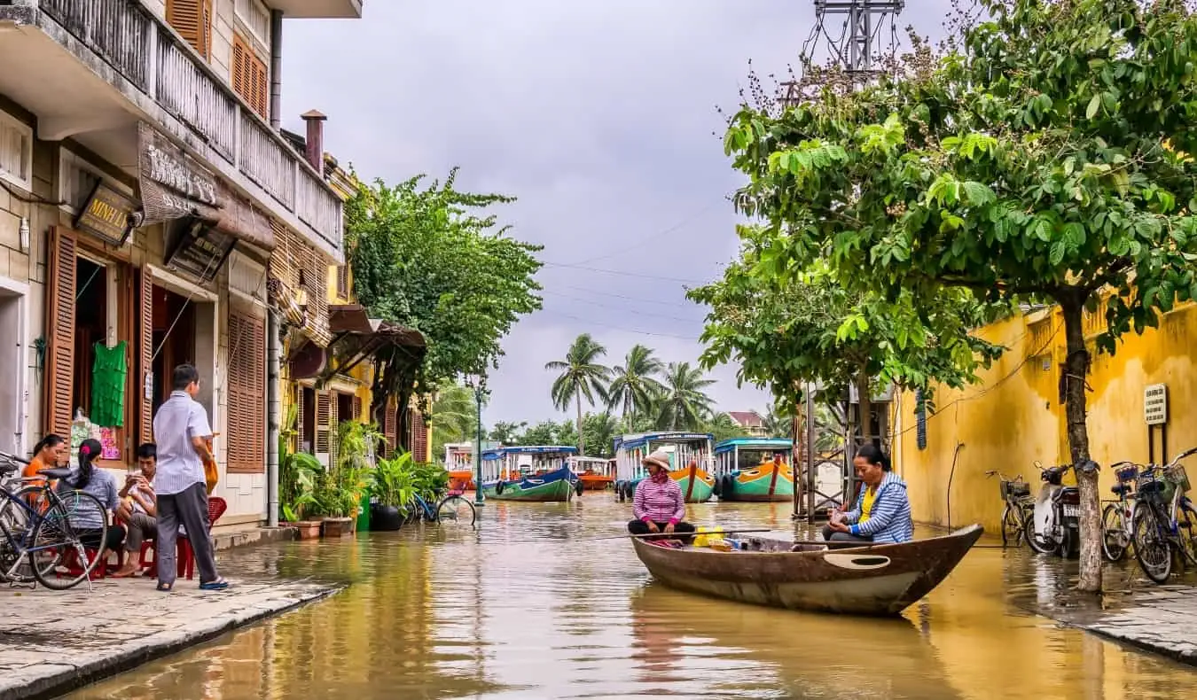 Kvinder sidder i en båd med folk på fortovet af bygninger i nærheden i Hoi An, Vietnam