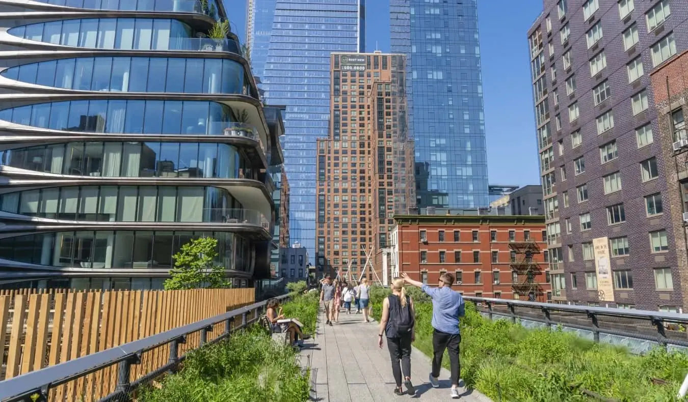 Orang yang berjalan di laluan yang dikelilingi oleh kehijauan dan bangunan pencakar langit yang tinggi di The High Line Park di Daerah Pembungkusan Daging di NYC