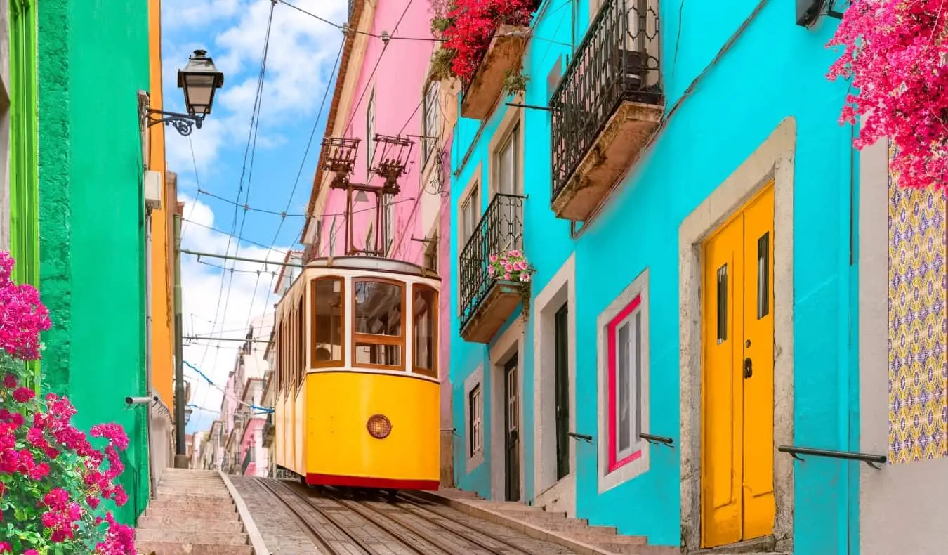 Historische gele tram op een straat die een heuvel afdaalt, omzoomd met kleurrijke gebouwen en bloemen op de balkons in Lissabon, Portugal