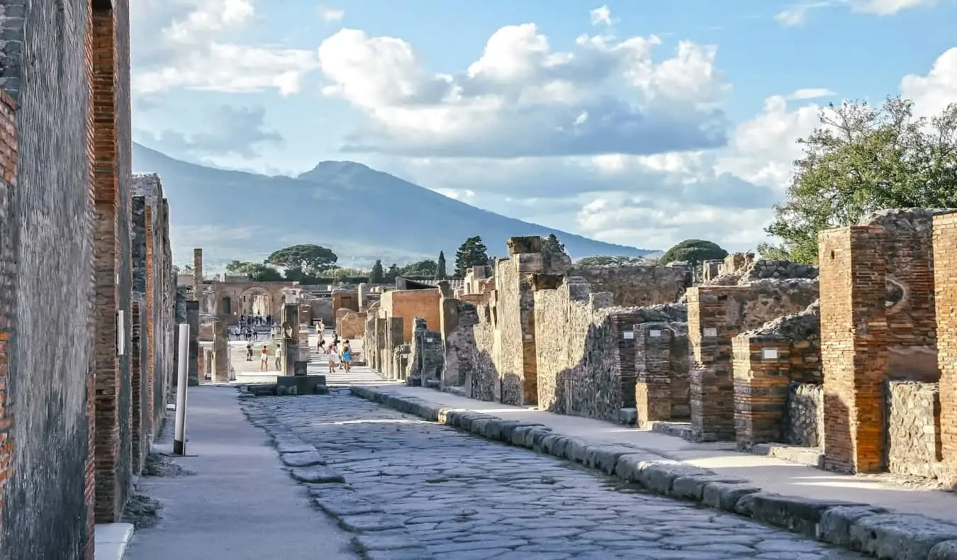 Calle adoquinada en Pompeya, bordeada de ruinas de edificios y el Monte Vesubio al fondo.