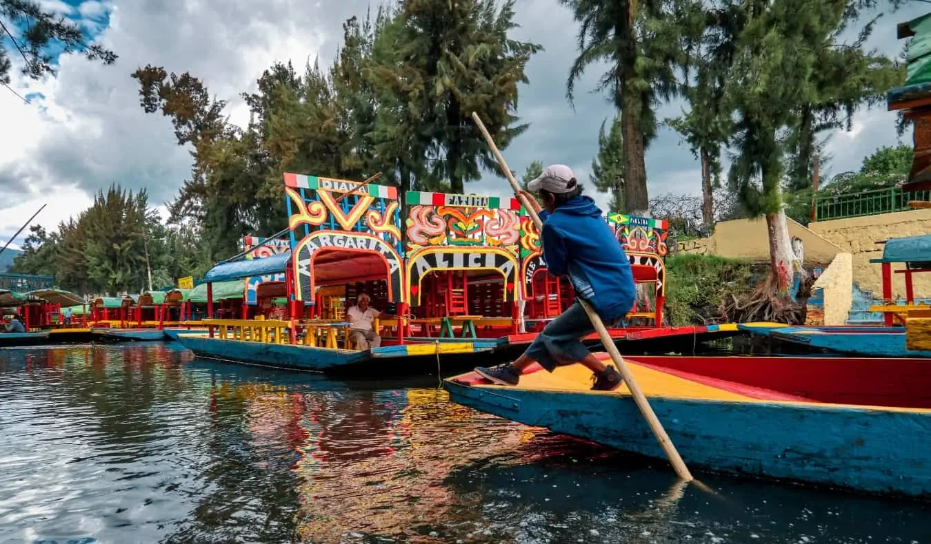 میکسیکو سٹی، میکسیکو میں Xochimilco نہروں میں ایک نوجوان لڑکا ایک لمبی چھڑی کے ساتھ رنگین کشتی کو دریا کے نیچے دھکیل رہا ہے۔