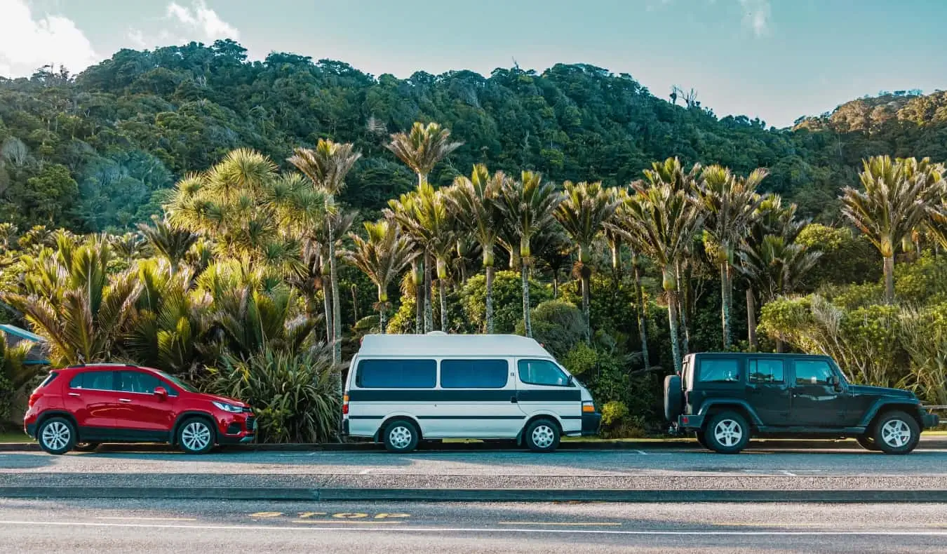 Sebuah kereta, campervan, dan Jeep di jalan raya di hutan hujan New Zealand.