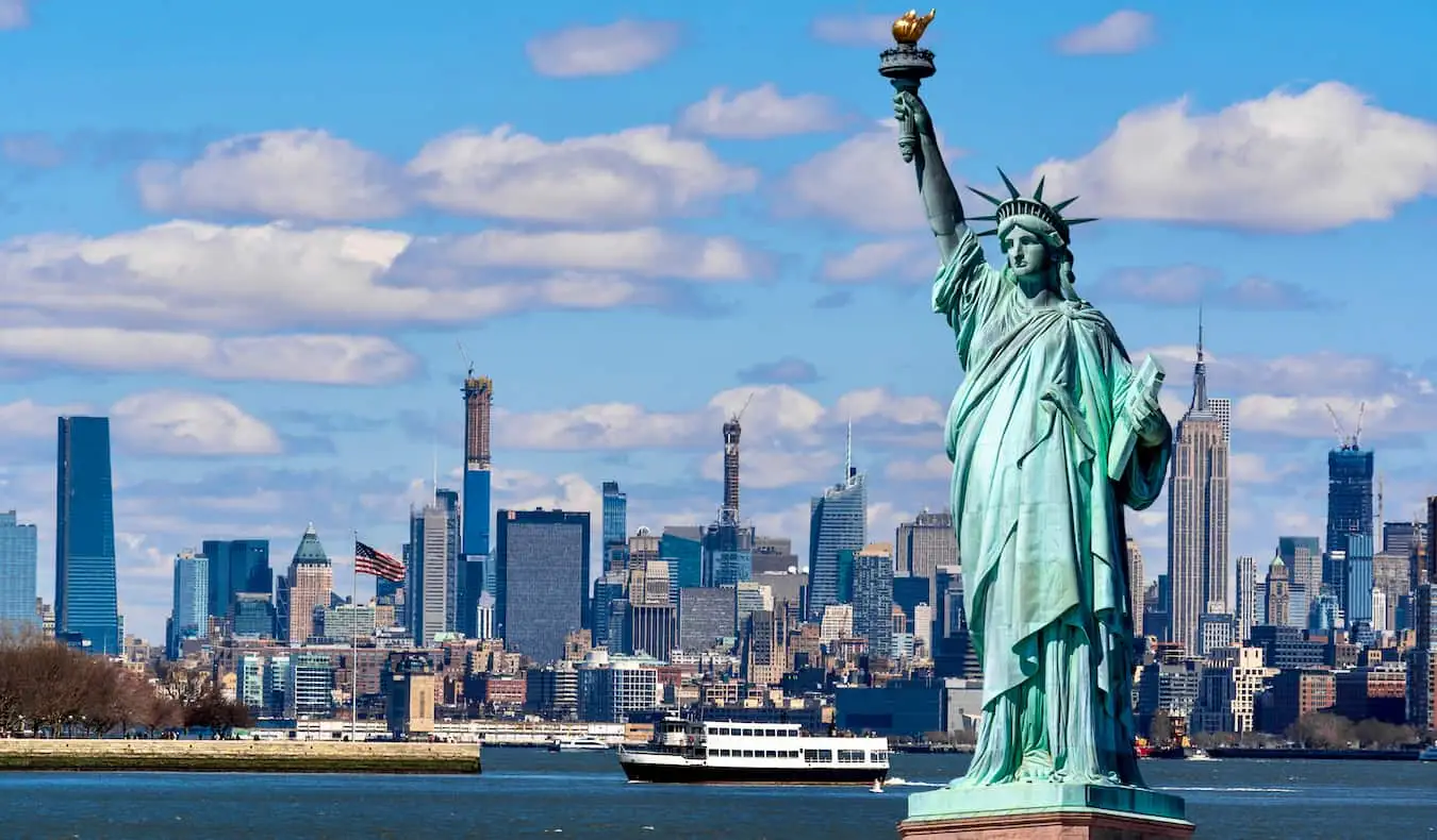 Die ikonische Freiheitsstatue mit NYC im Hintergrund an einem sonnigen Tag mit blauem Himmel