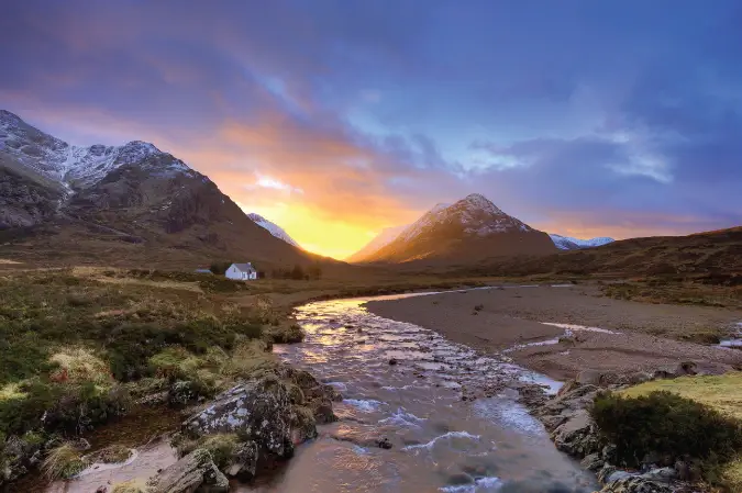 Foto matahari terbenam di balik pegunungan di Glencoe, Skotlandia dengan sebuah rumah di tengahnya