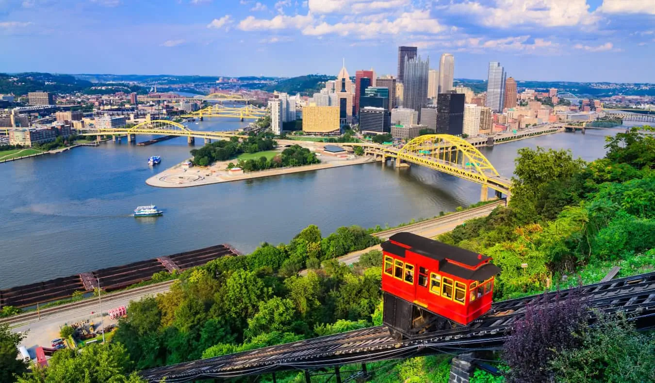 Een rode kabelbaan die op de voorgrond de berg op gaat met de skyline van Pittsburgh, PA met zijn vele bruggen over de rivier op de achtergrond