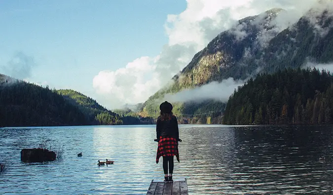 isang babaeng nakatayo sa dulo ng isang pantalan kung saan matatanaw ang isang alpine lake