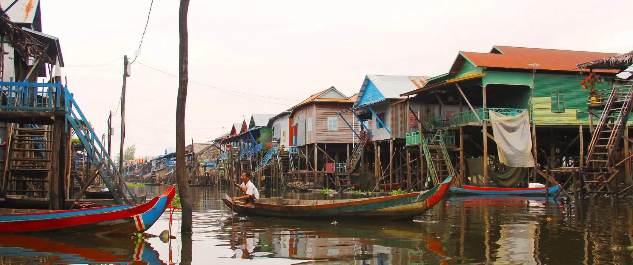 En man som använder en stolpe för att manövrera en lång båt nerför en vattenväg omgiven av färgglada hus på pålar i Tonle Sap, Kambodja