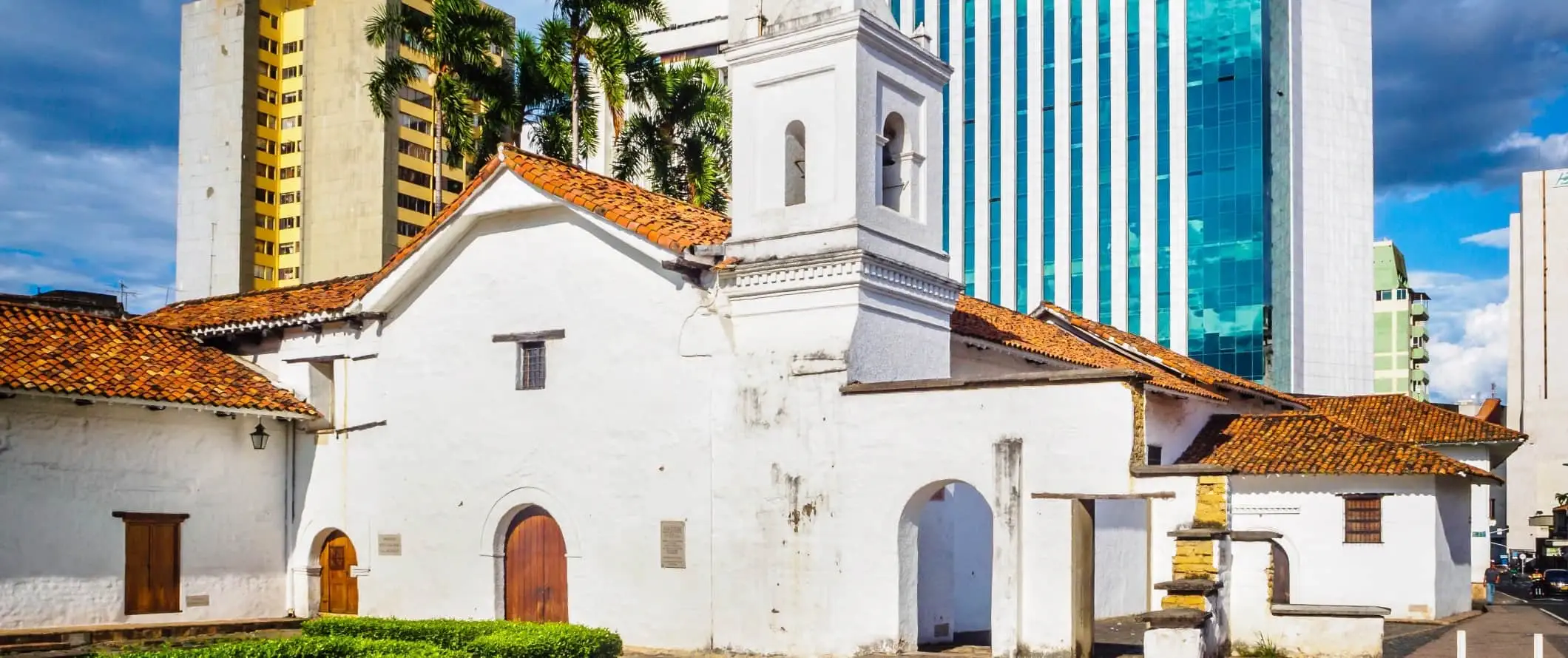 Het Museum voor Religieuze Kunst La Merced, een witgekalkt 17e-eeuws gebouw met een rood terracotta dak tegen moderne wolkenkrabbers in Cali, Colombia