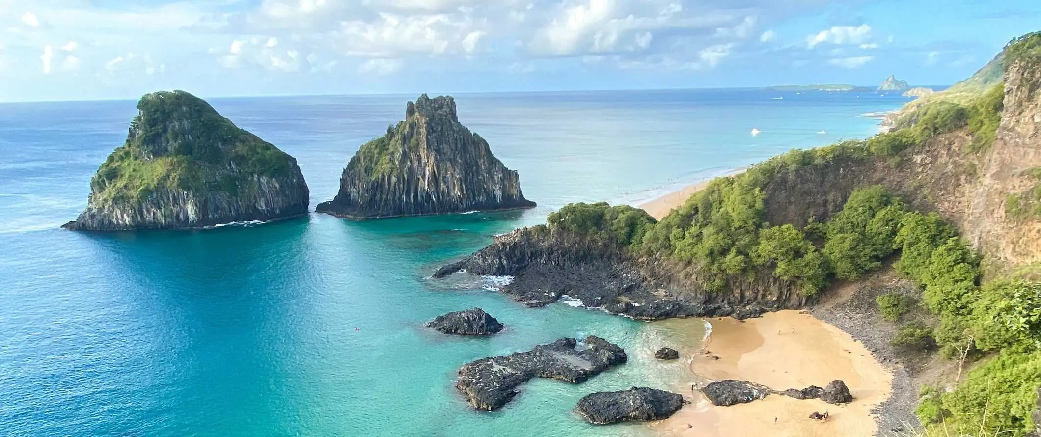 Pemandangan panorama pantai dan dua formasi batu besar yang muncul dari perairan pirus Fernando de Noronha, Brasil