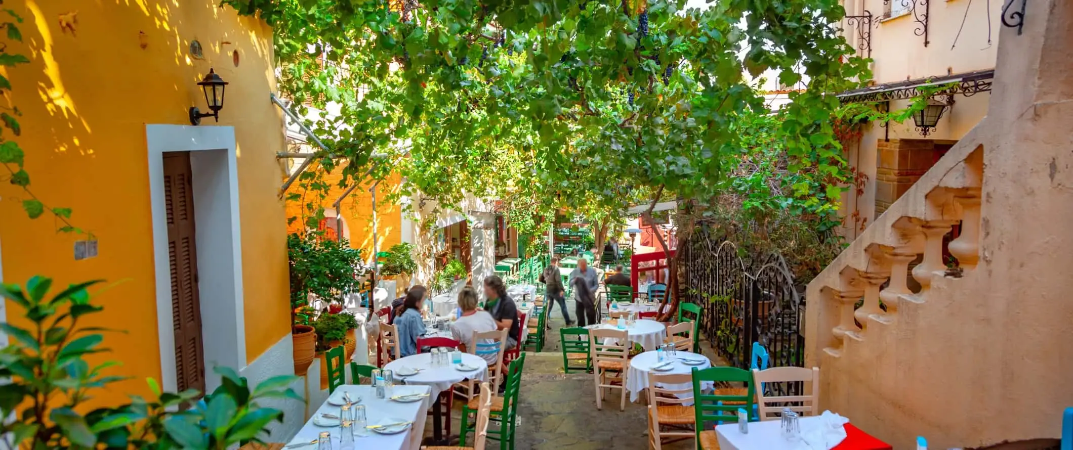 Tables de café bordant une ruelle couverte de branches d'arbres luxuriantes, entourées de bâtiments jaune vif, à Athènes, en Grèce.