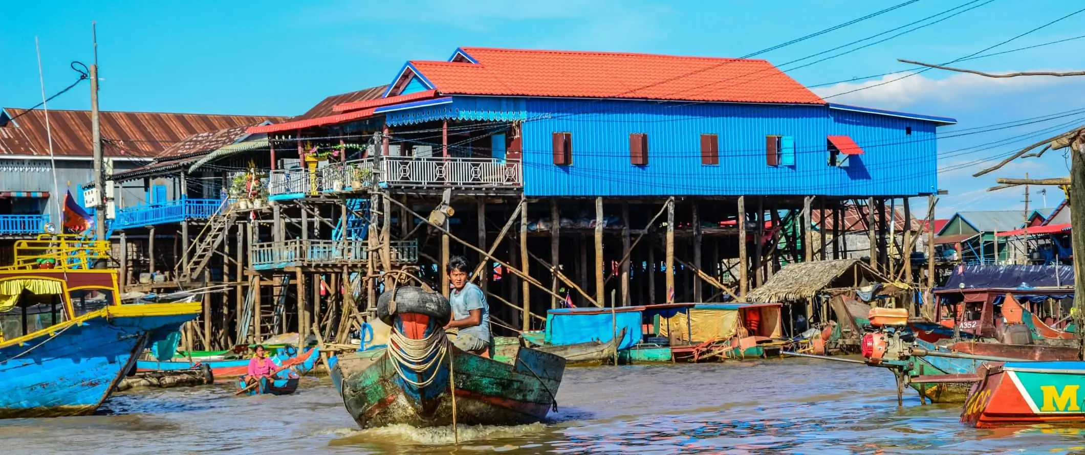 Man bestuurt een boot over een waterweg voor felgekleurde huizen op palen in Tonle Sap, Cambodja