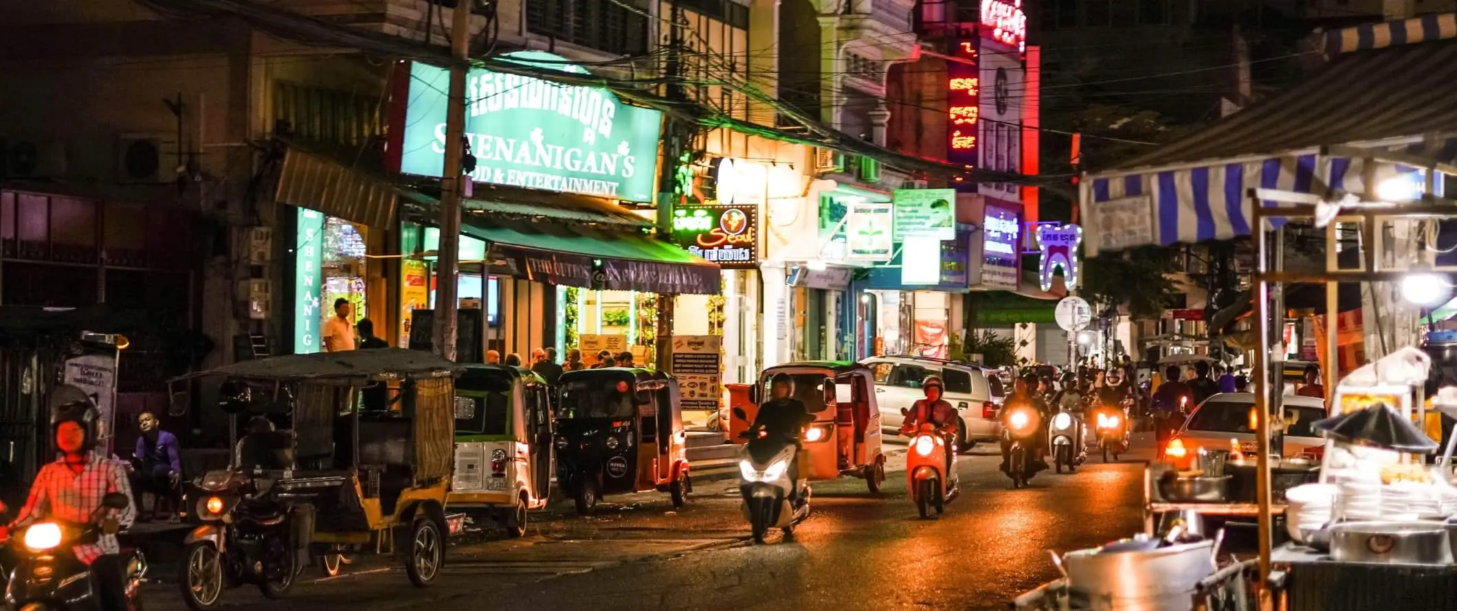 Bromfietsen, motorfietsen en tuk-tuks rijden door een straat die 's nachts verlicht is in Phnom Penh, de hoofdstad van Cambodja