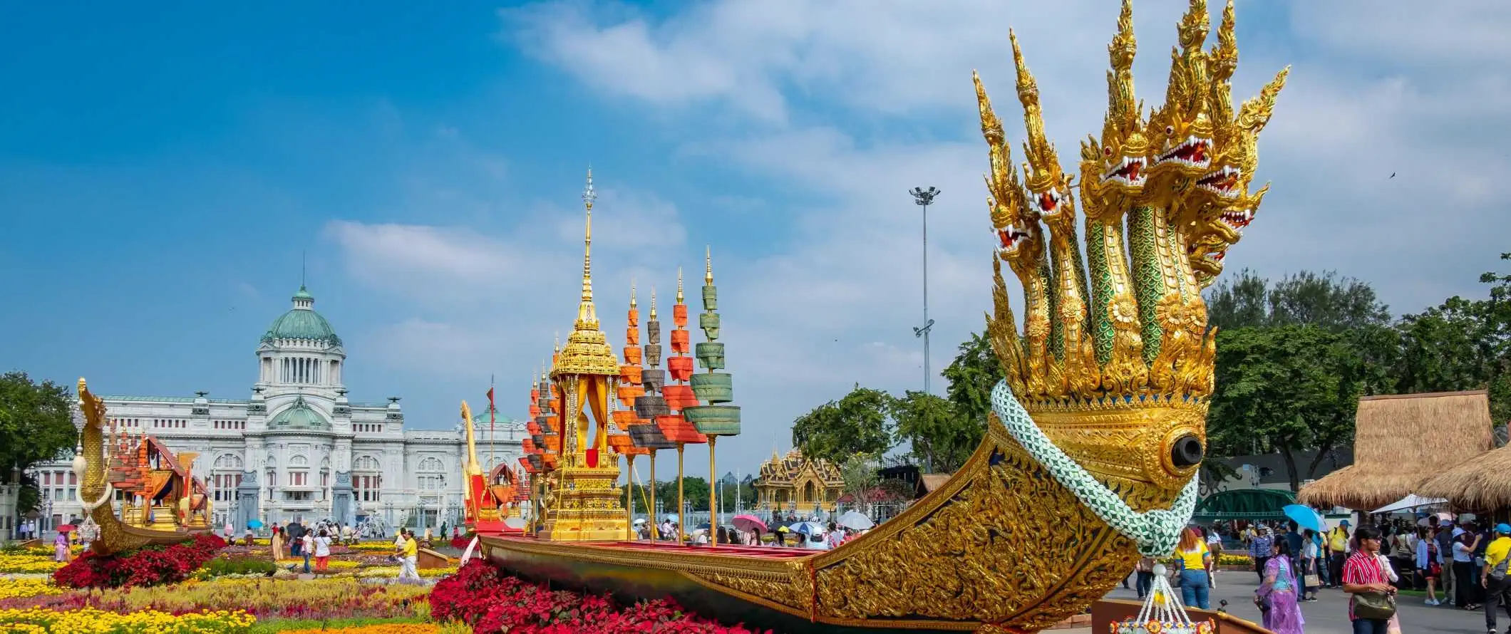 En stor, lang gylden pram med mange dragehoveder sidder på et lyst blomsterbed foran Royal Barge Museum i Bangkok, Thailand