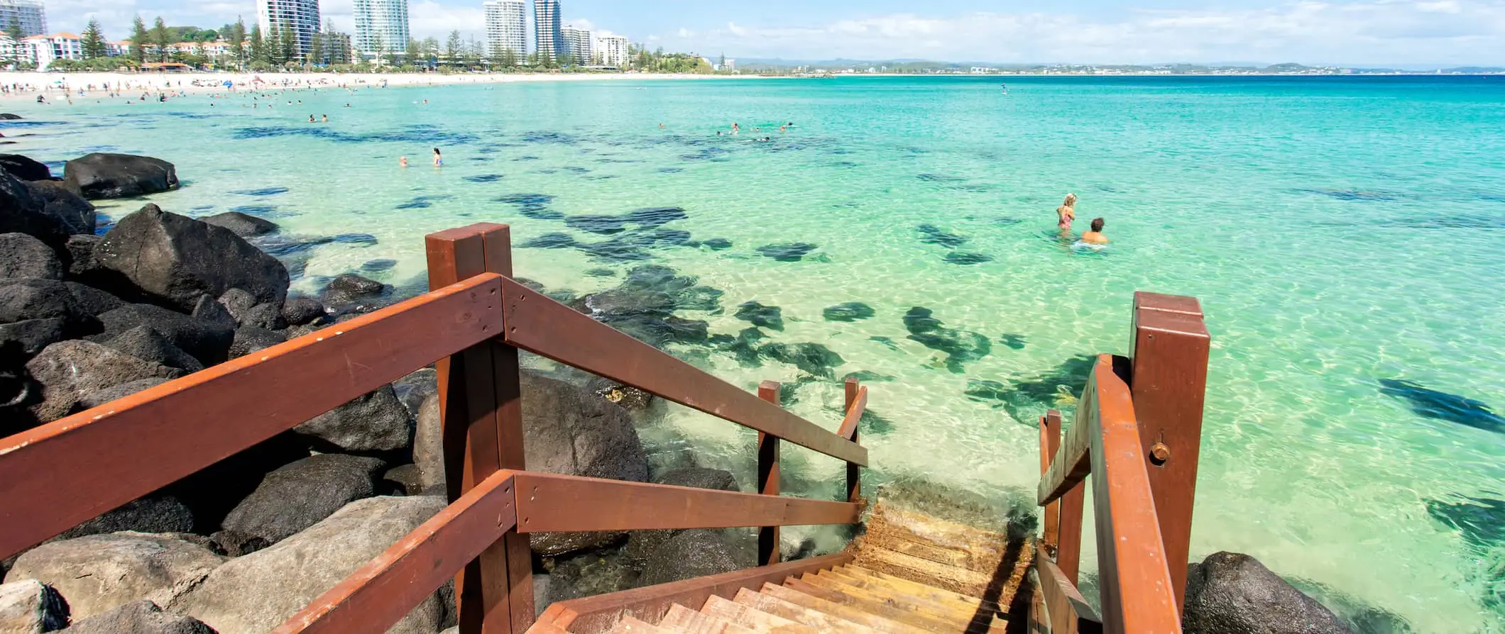 Menschen schwimmen im klaren Wasser der Gold Coast, Australien, in der Nähe einer kleinen Holztreppe mit der Stadt im Hintergrund