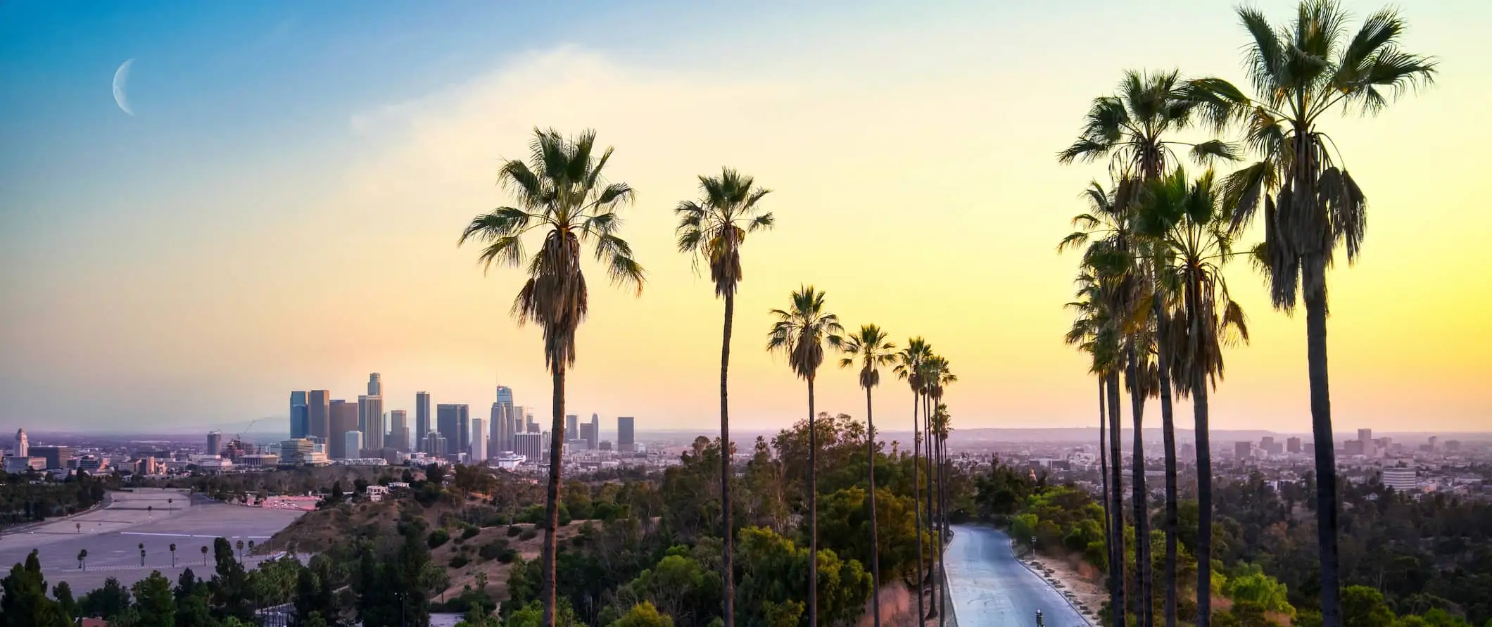 Los Angeles auringonlaskun aikaan, taustalla kohoavia rakennuksia ja palmuja etualalla