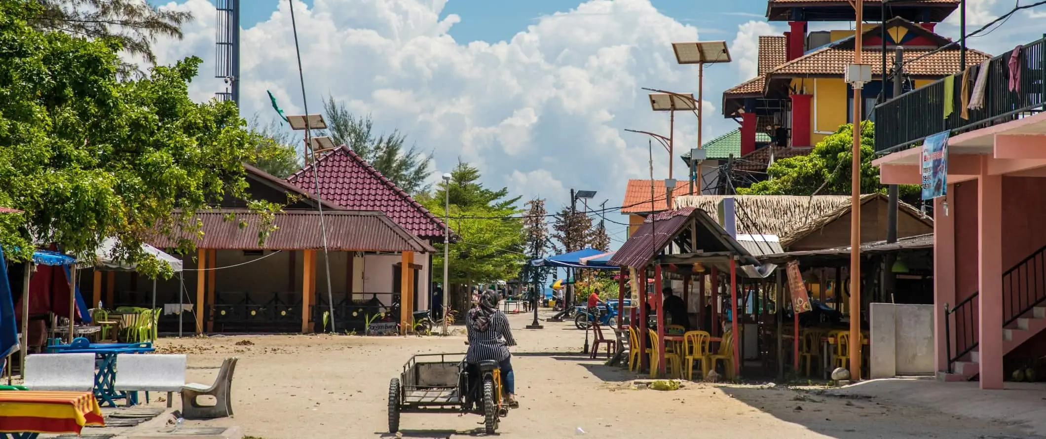 En person som åker motorcykel på en sandgata med låga byggnader på båda sidor i Perhentian Islands, Malaysia